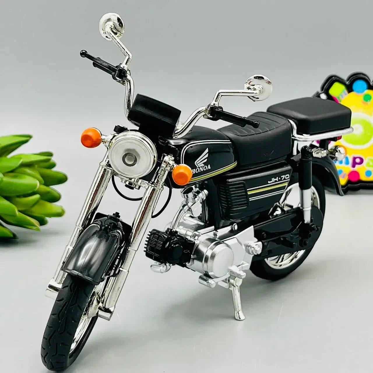 A detailed miniature model of a black Honda JH-70 motorcycle with chrome accents is displayed against a gray background. The model showcases the vintage design, complete with a headlight, orange indicator lights, and a black seat.