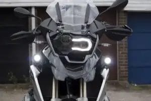 Front view of a gray BMW motorcycle, highlighting its distinctive headlight design, windscreen, handguards, and auxiliary lights. The bike is parked in front of a brick garage door.